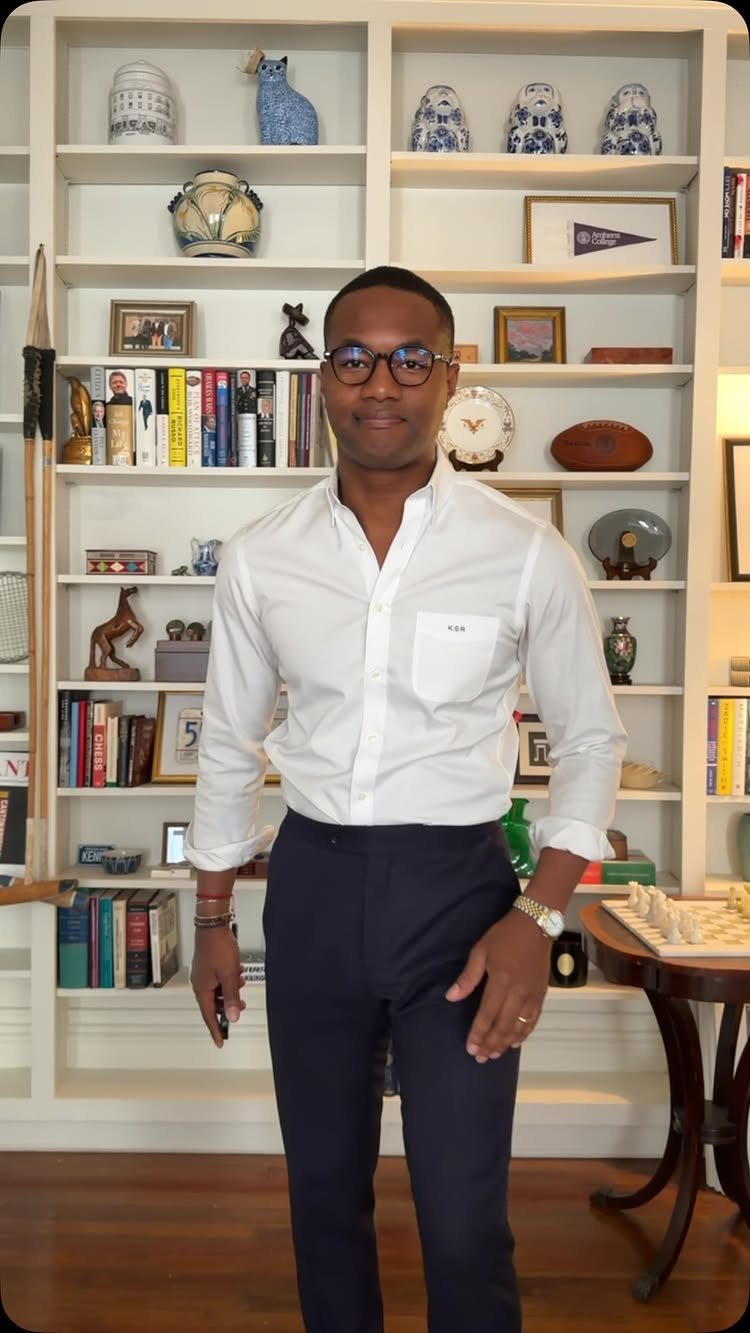 Man in white button-down shirt and navy trousers standing before a white bookshelf.