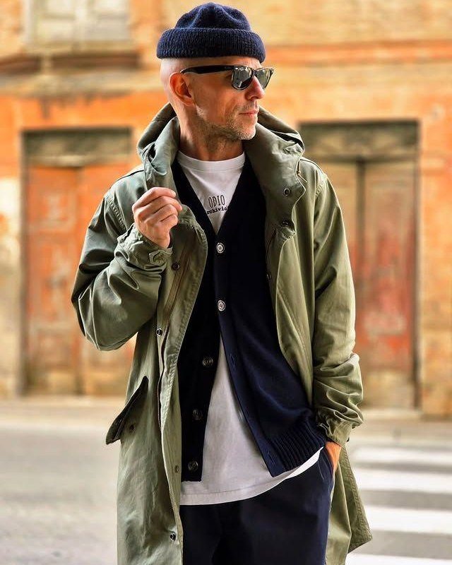 Man wearing an olive green hooded parka, navy cardigan, and navy beanie outdoors.