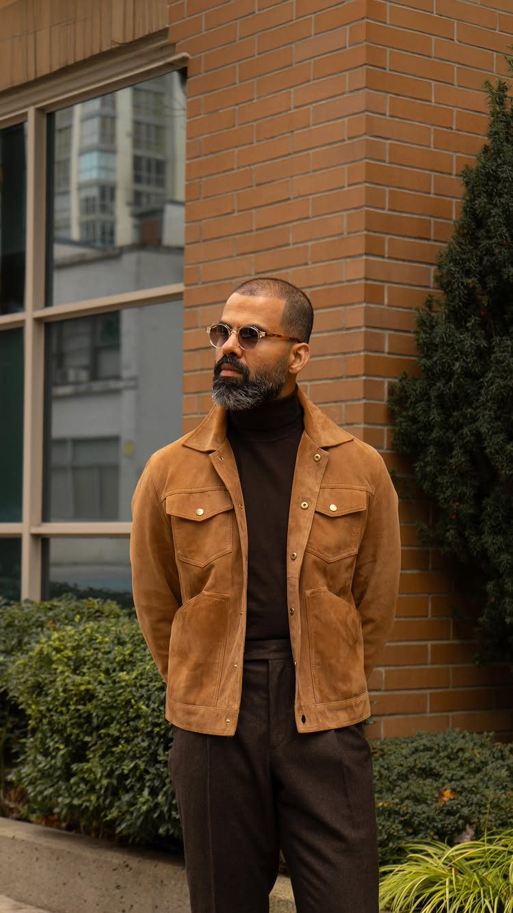 Man wearing a tan suede trucker jacket and dark brown turtleneck against brick wall