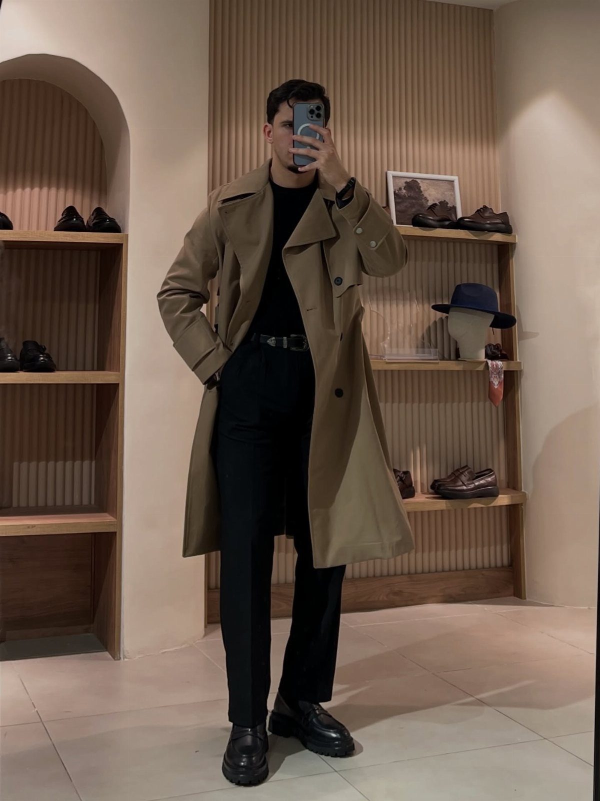 Man wearing a tan trench coat and black trousers in a shoe store.