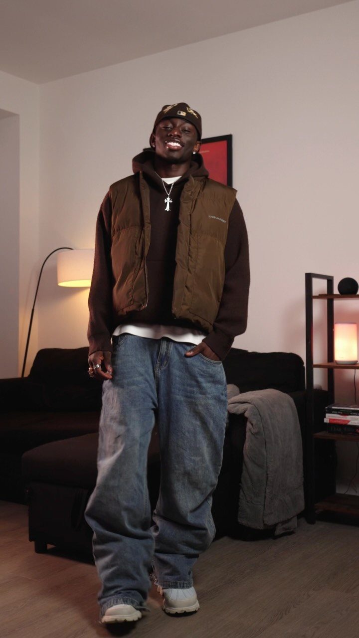 Man wearing a brown puffer vest over a brown hoodie and baggy blue jeans.