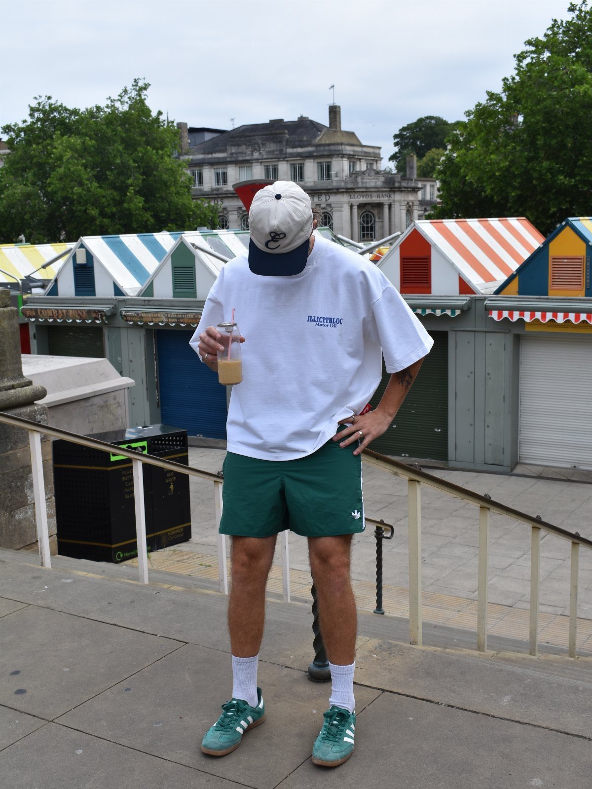 Person wearing green cotton shorts, white t-shirt, and green sneakers by beach huts.