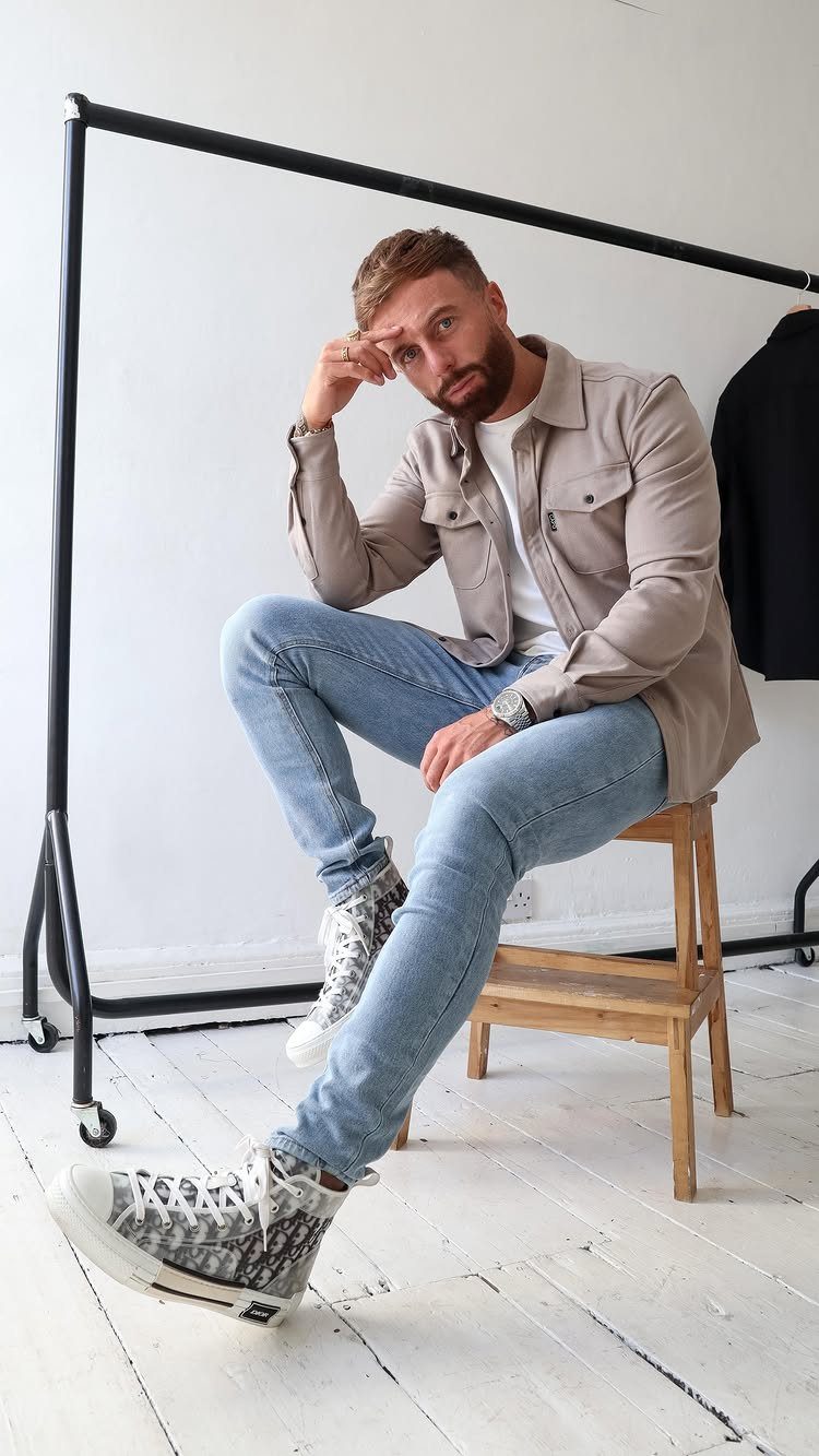 Man sitting on a stool wearing a beige overshirt and light blue jeans.