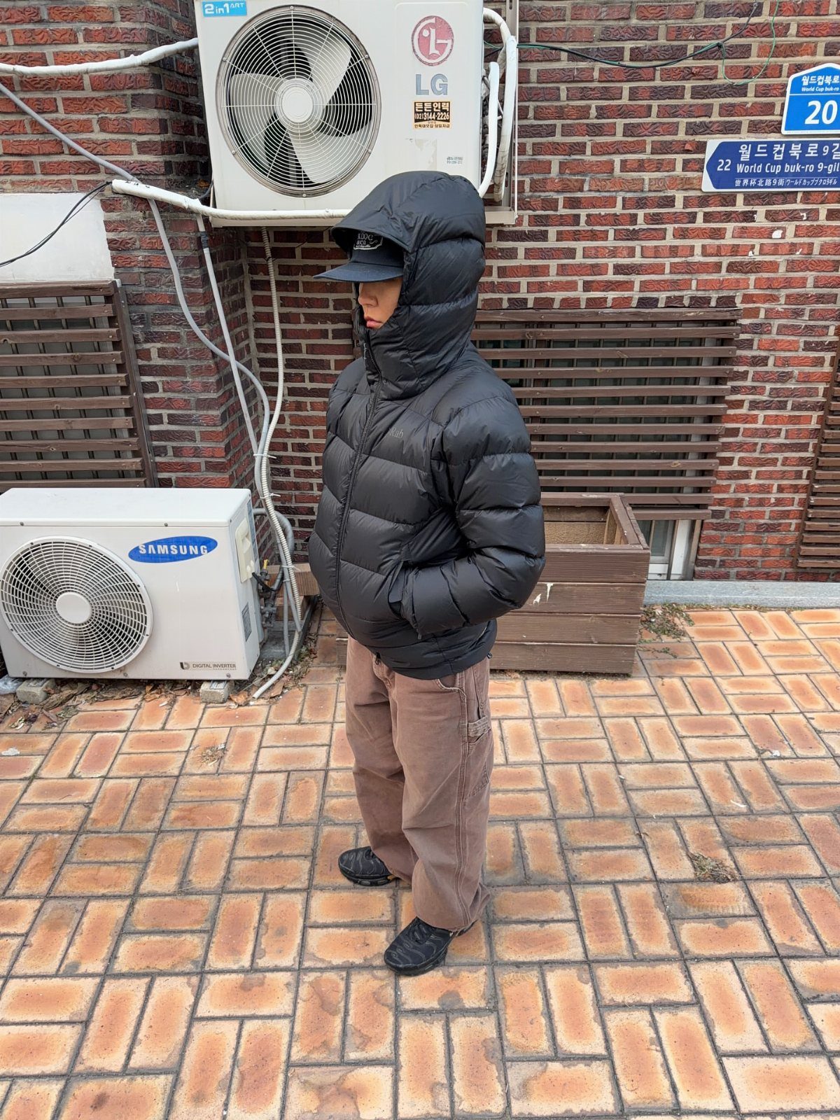 Person wearing a black hooded puffer jacket and brown trousers on brick pavement.