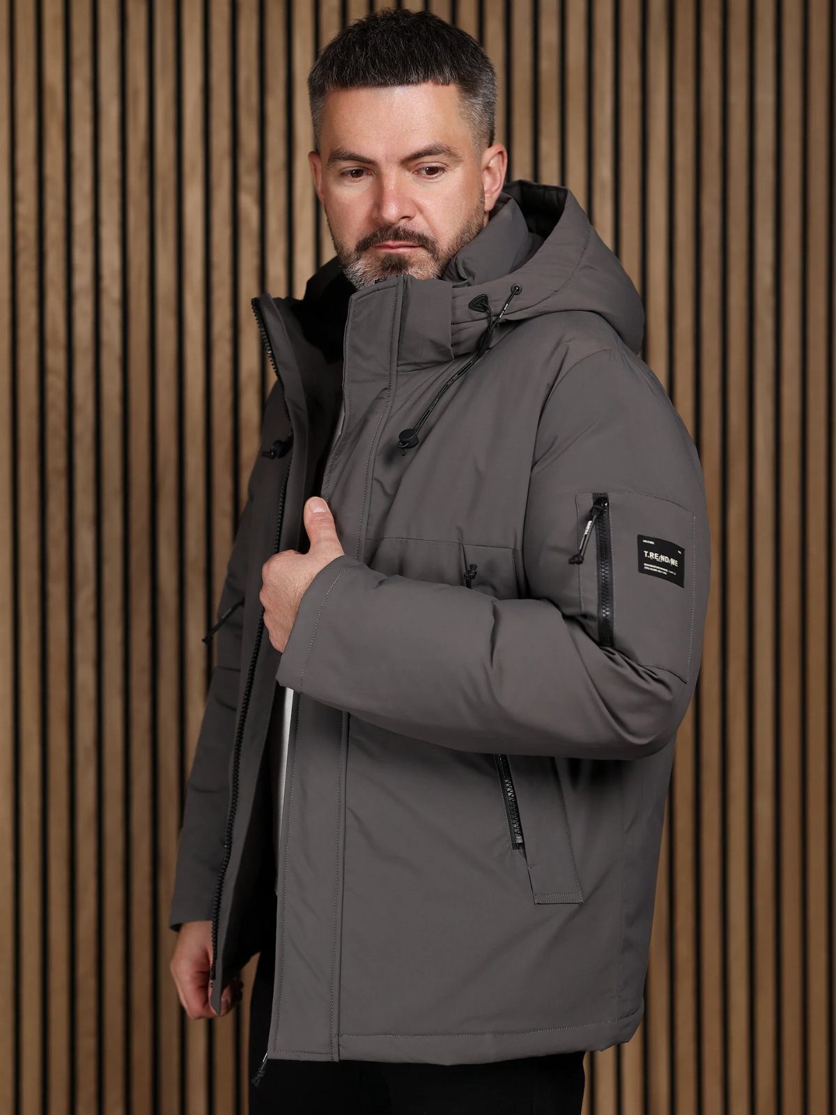 Man wearing a grey hooded winter jacket against a wooden slat wall background.