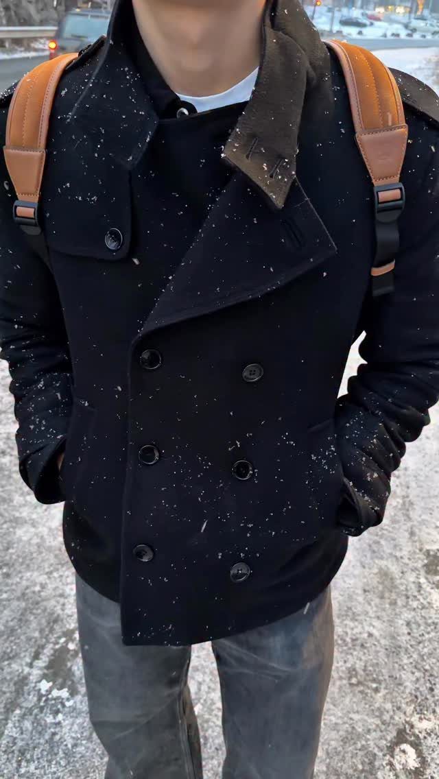 Person wearing a black double-breasted pea coat and brown backpack on snowy ground