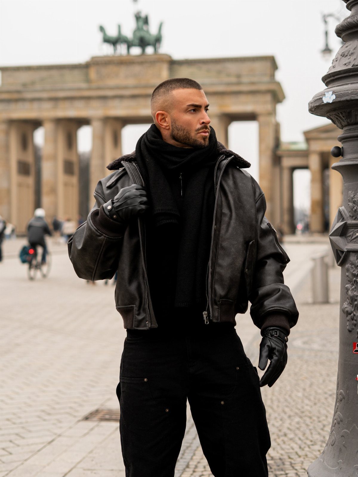Man in black leather jacket and scarf standing before the Brandenburg Gate.