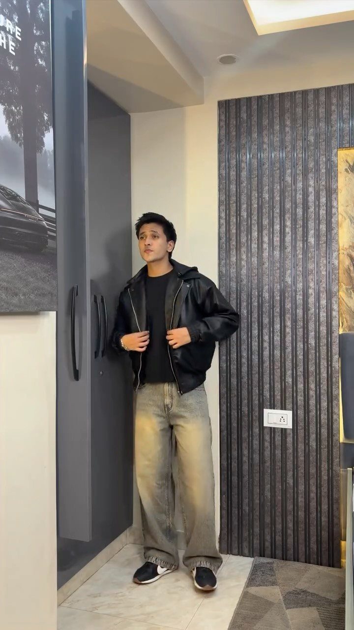 Man wearing a black leather jacket and wide-leg gray jeans standing indoors.