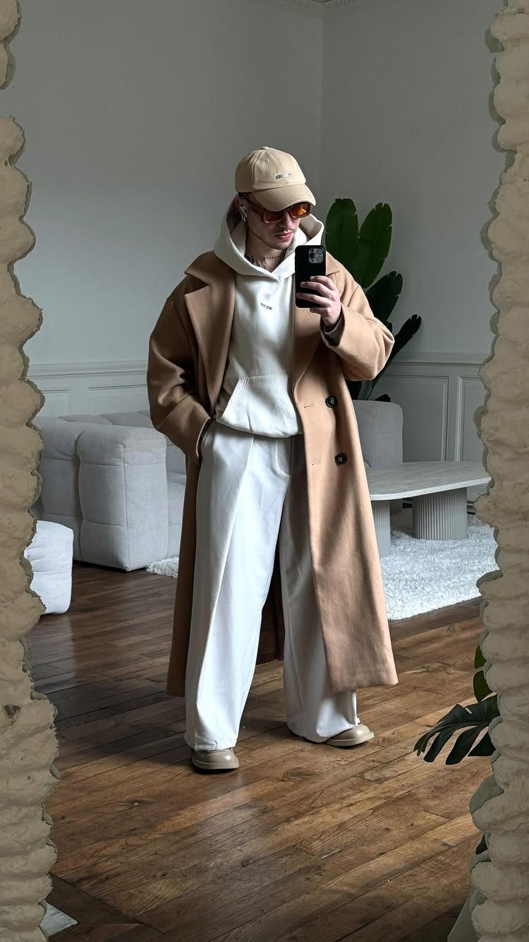 Person in a long tan coat and wide-leg white trousers taking a mirror selfie.