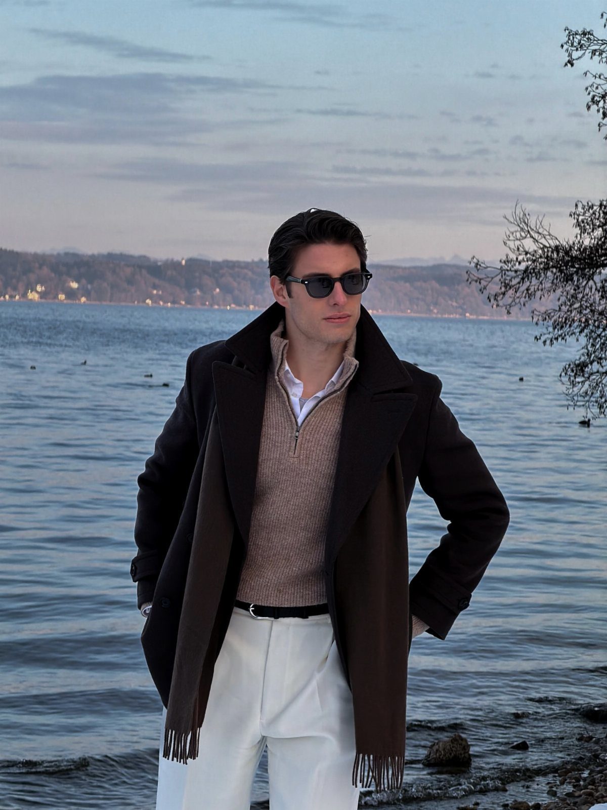 Man in brown overcoat and beige sweater standing by a lake at dusk.