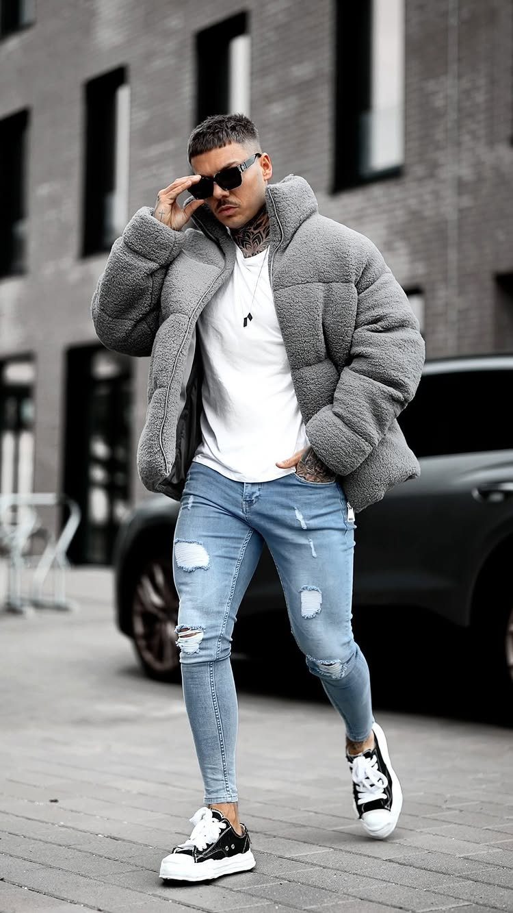 Man in grey puffer jacket and ripped blue jeans posing on a city street