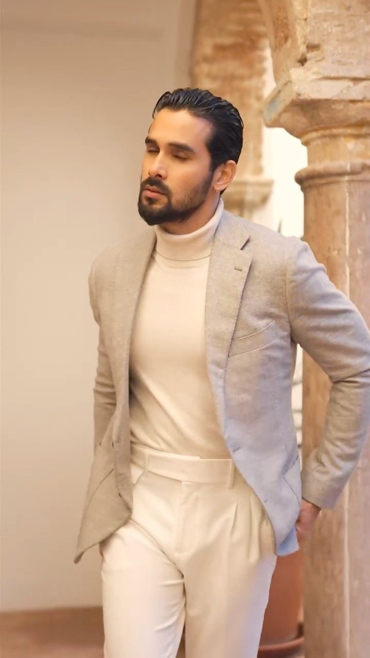 Man wearing a light gray blazer, cream turtleneck, and white trousers by a pillar.