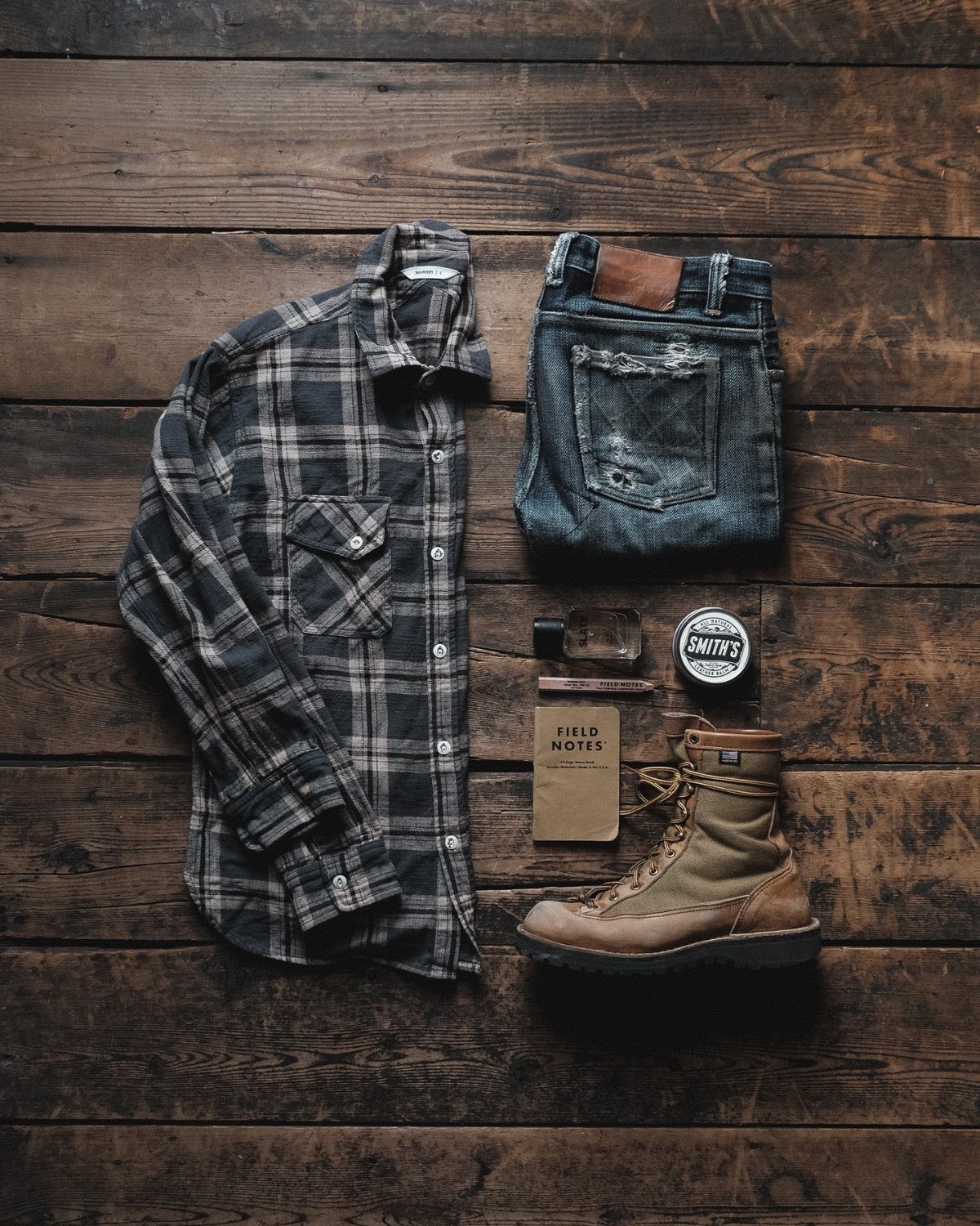 Grey plaid flannel shirt, blue jeans, and brown boot arranged on wooden floor.