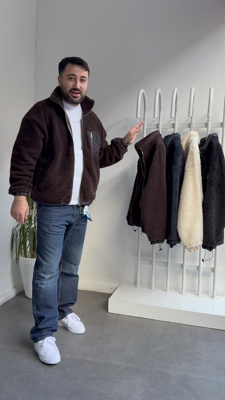 Man wearing a brown fleece jacket and blue jeans standing by a clothing rack