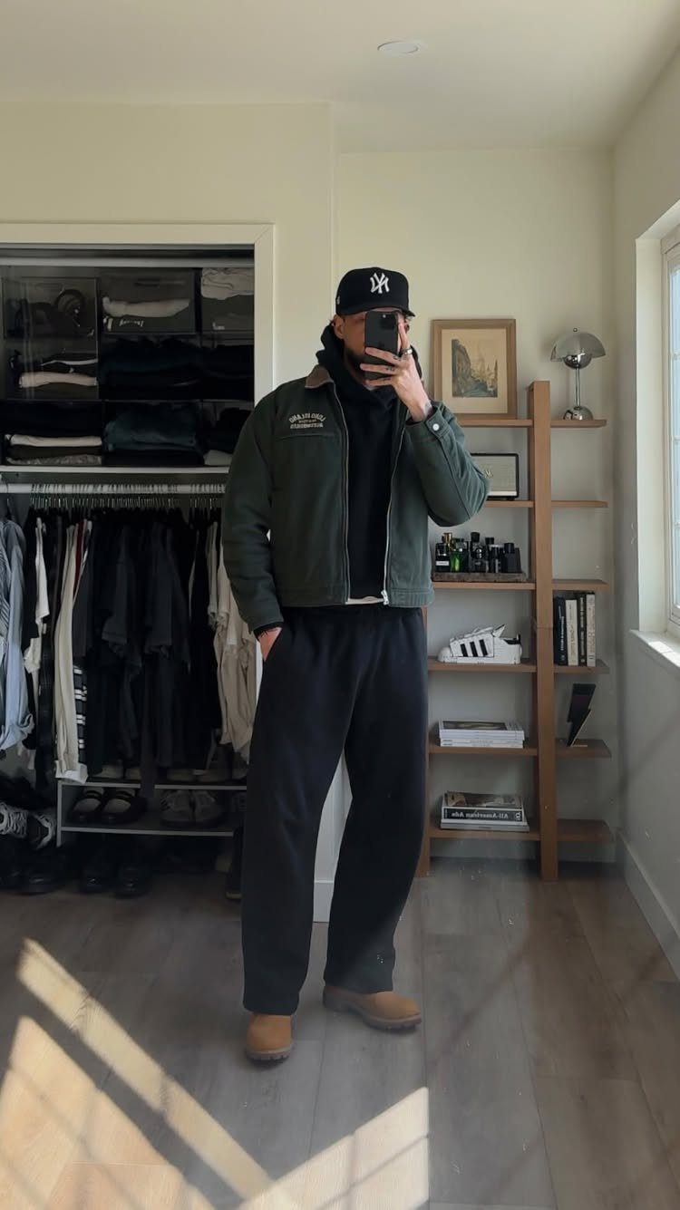 Man wearing a green jacket, black hoodie, wide-leg trousers, and a baseball cap.