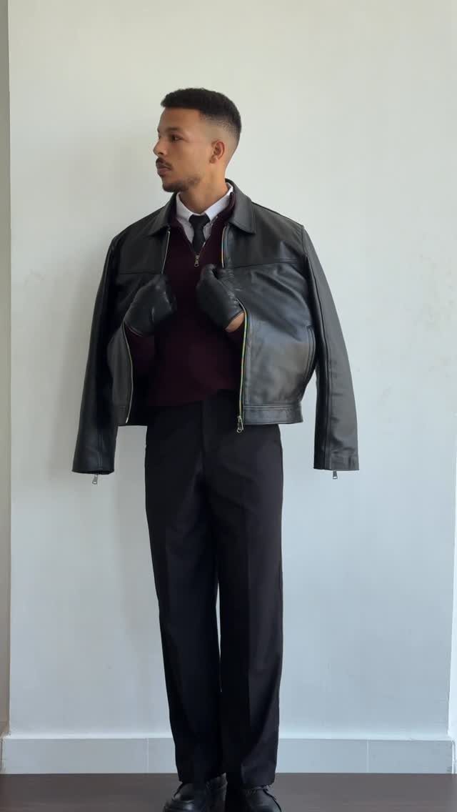 Man wearing a leather jacket, burgundy sweater, tie, and black trousers against white wall