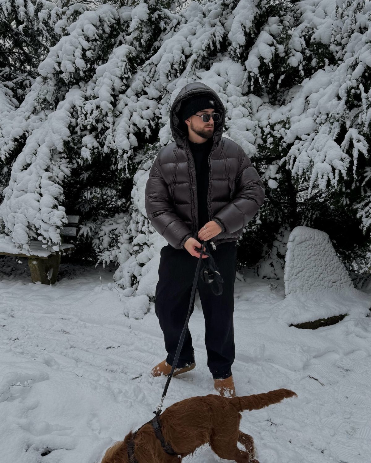Man in a grey puffer jacket and black pants standing in snowy woods with dog