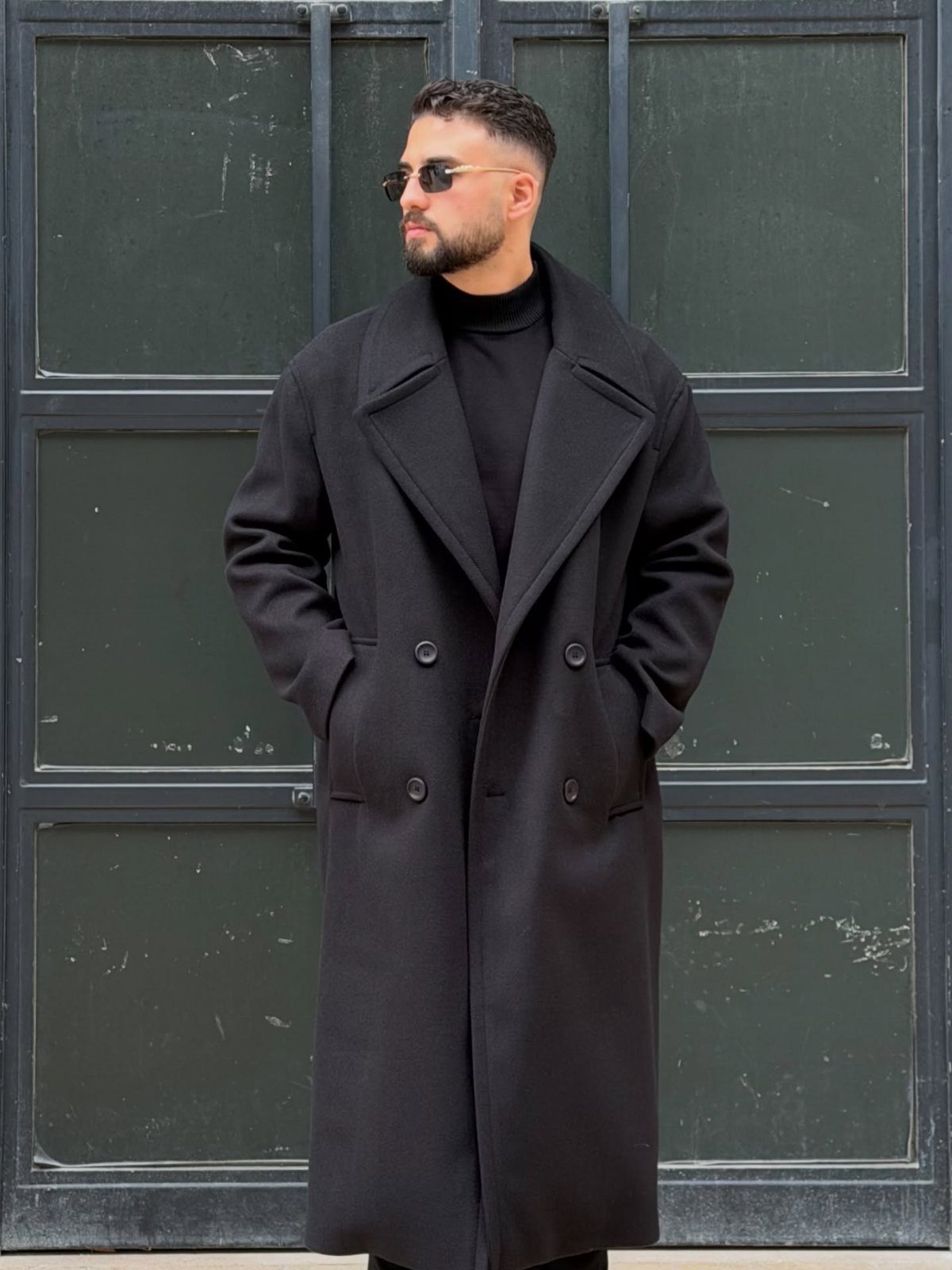 Man in a black wool overcoat and black turtleneck standing by a door