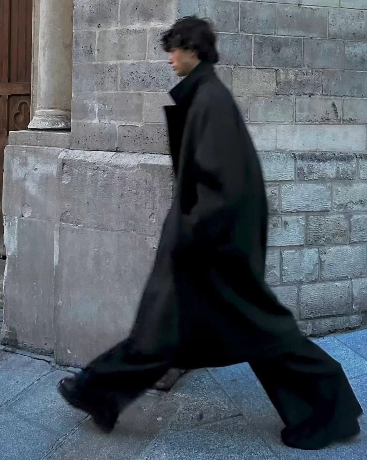 Person in an oversized black wool overcoat walking past a stone wall.