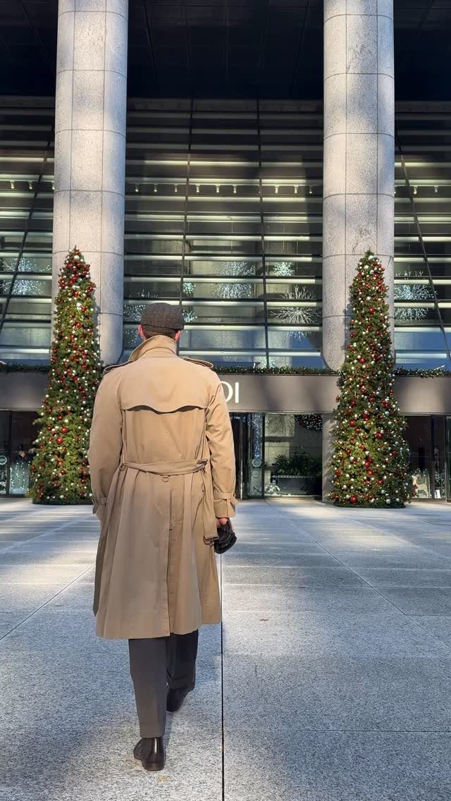 Man in a tan belted trench coat walking toward decorated Christmas trees.