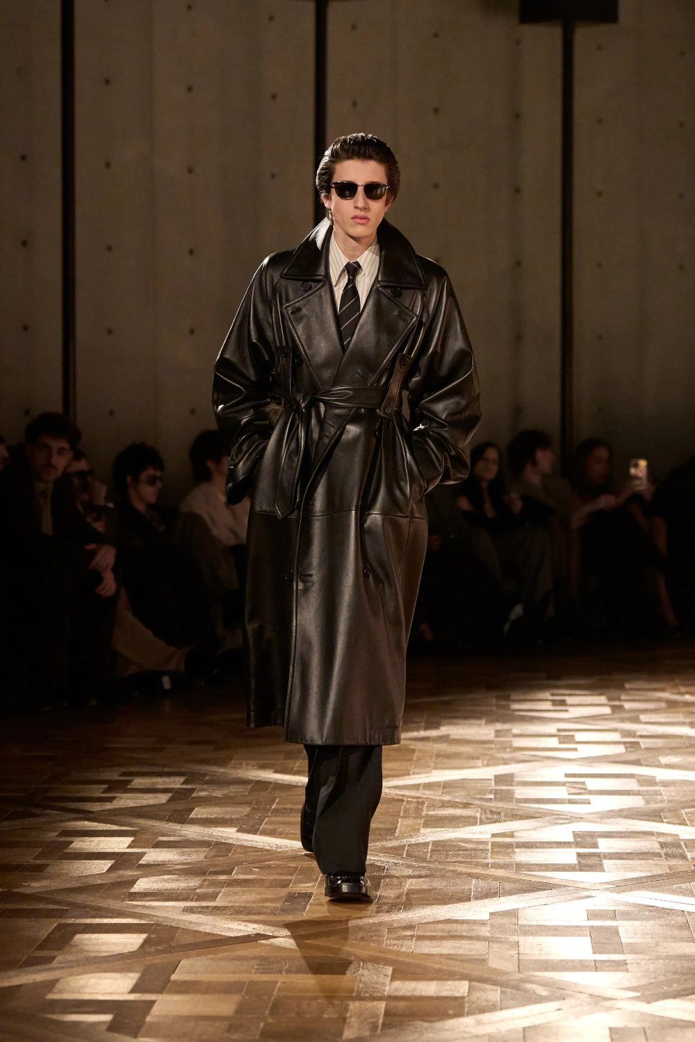 Male model wearing a black leather trench coat walking on a wooden runway