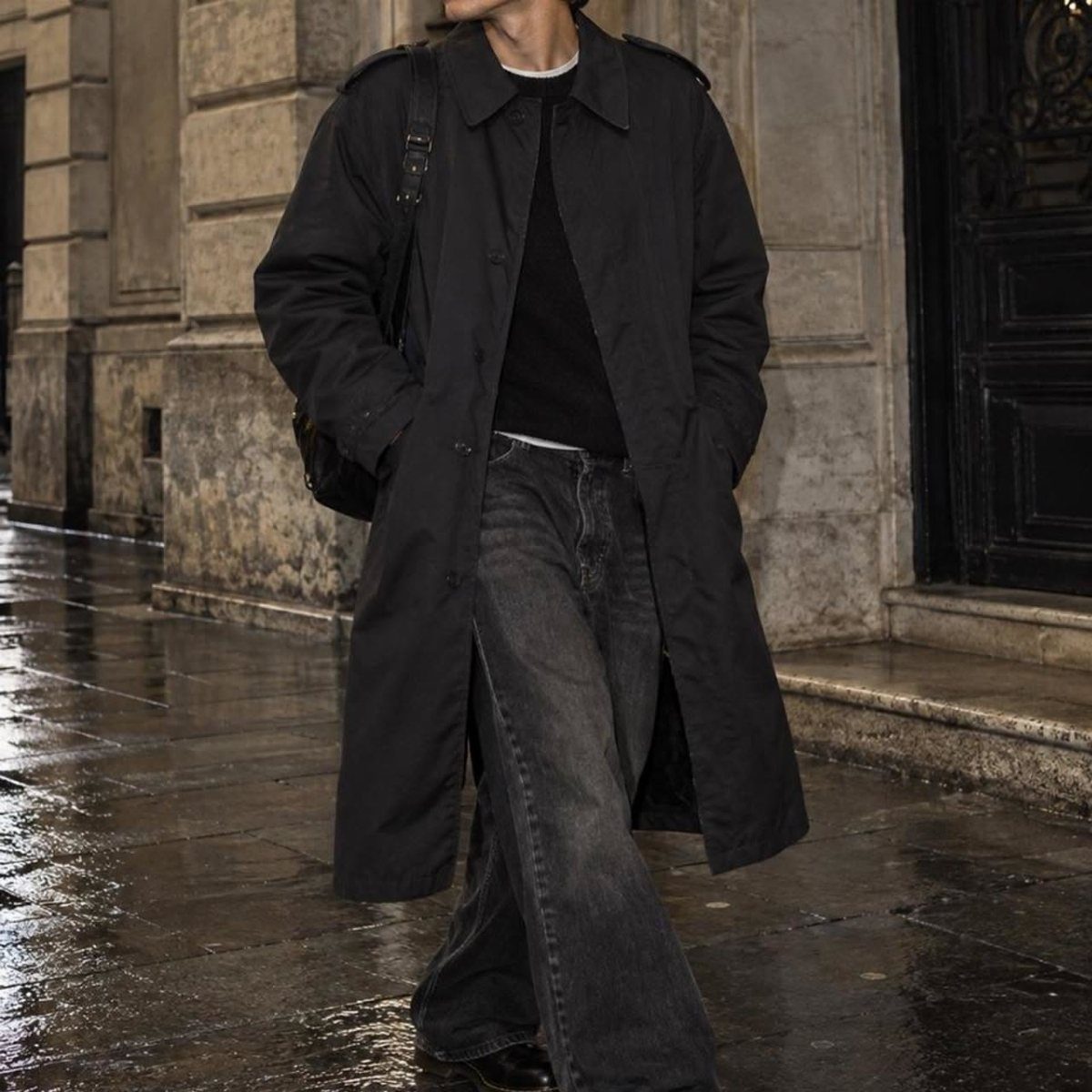 Person wearing a long black trench coat and grey jeans on wet street